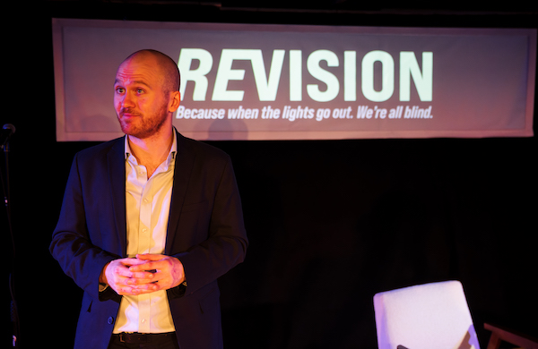 Tim, played by Samuel Brewer, giving a presentation in front of a screen that says ReVision. Because when the lights go out. We're all blind